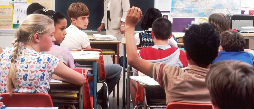 A classroom filled with children eagerly raising their hands to answer questions from the teacher.