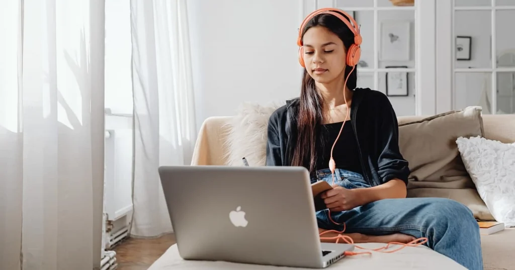 A young woman wearing orange headphones sits on a couch with a notebook, watching a laptop screen. Sunlight streams through a window, creating a cozy, relaxed atmosphere.