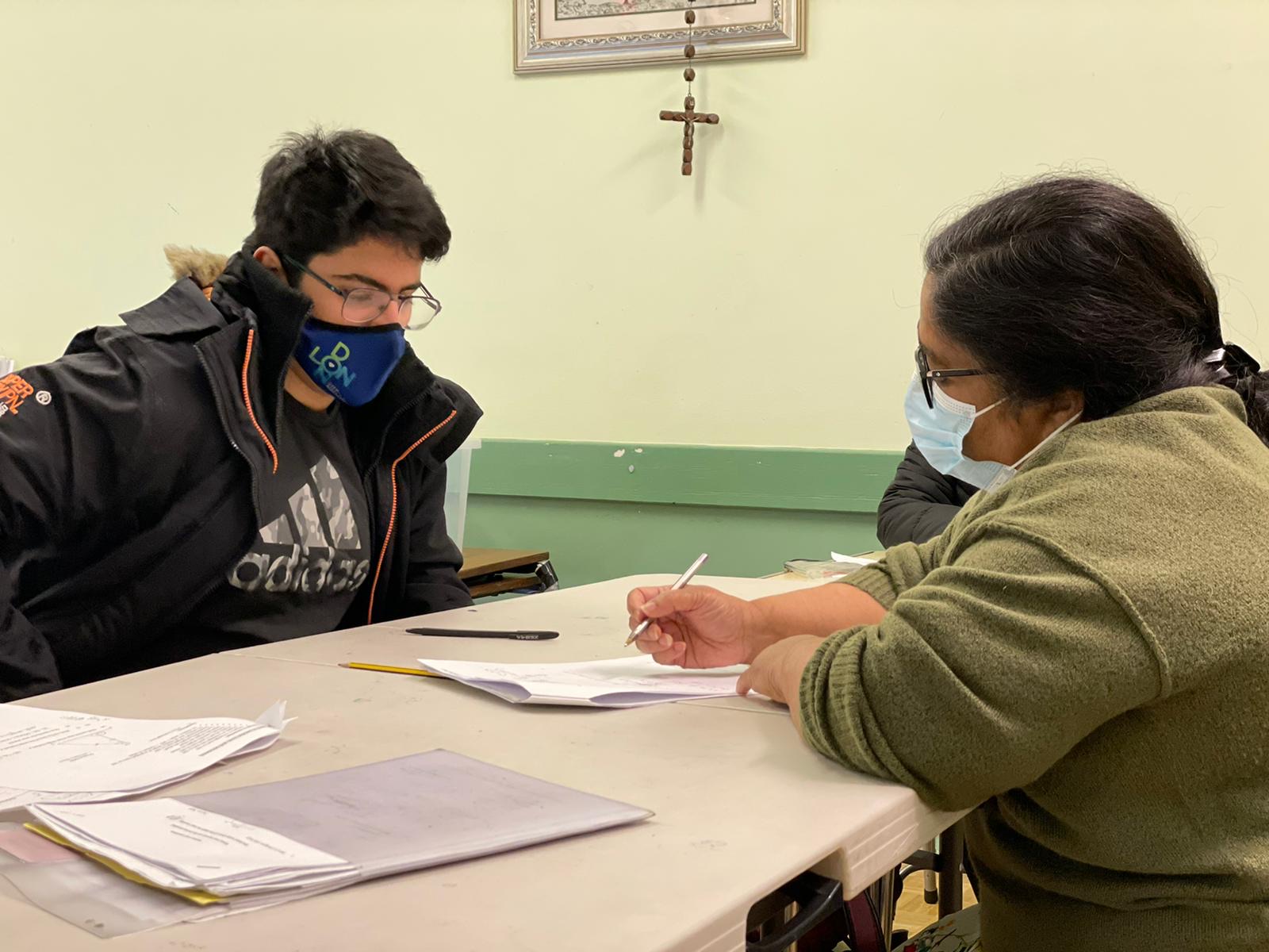 Two people wearing masks sit at a table covered with papers. One writes while the other watches attentively. A crucifix hangs on the wall behind them. Both appear focused on their work.