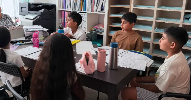 Four children sit around a table with papers, water bottles, and school supplies, facing an adult who appears to be leading a lesson in a classroom setting with shelves in the background.