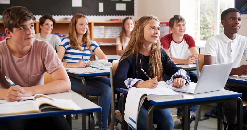 Students sit at desks in a classroom, taking notes and looking forward, with books and a laptop on their desks.